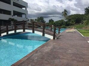 A swimming pool with a wooden bridge, a clear blue sky, and surrounding greenery.