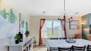 Un'area pranzo con un tavolo apparecchiato per quattro persone, un lampadario e vista su un paesaggio verde attraverso una finestra con tende rosse.