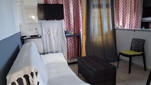 A room with a bed, a television, a small table, a chair, and a window with curtains. - APARTMENT LA DAME DE FER (GOSIER)