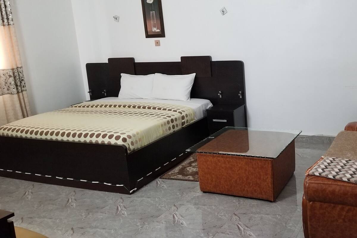 A bedroom with a bed, a wooden nightstand, a brown ottoman, and a brown leather sofa.