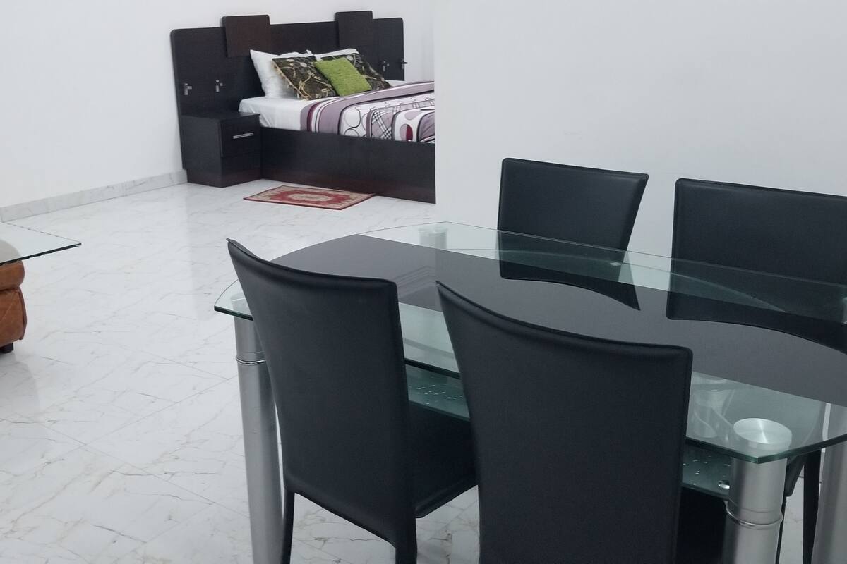 A modern dining area with a glass table and black chairs, a sofa, and a bed with a headboard in the background.