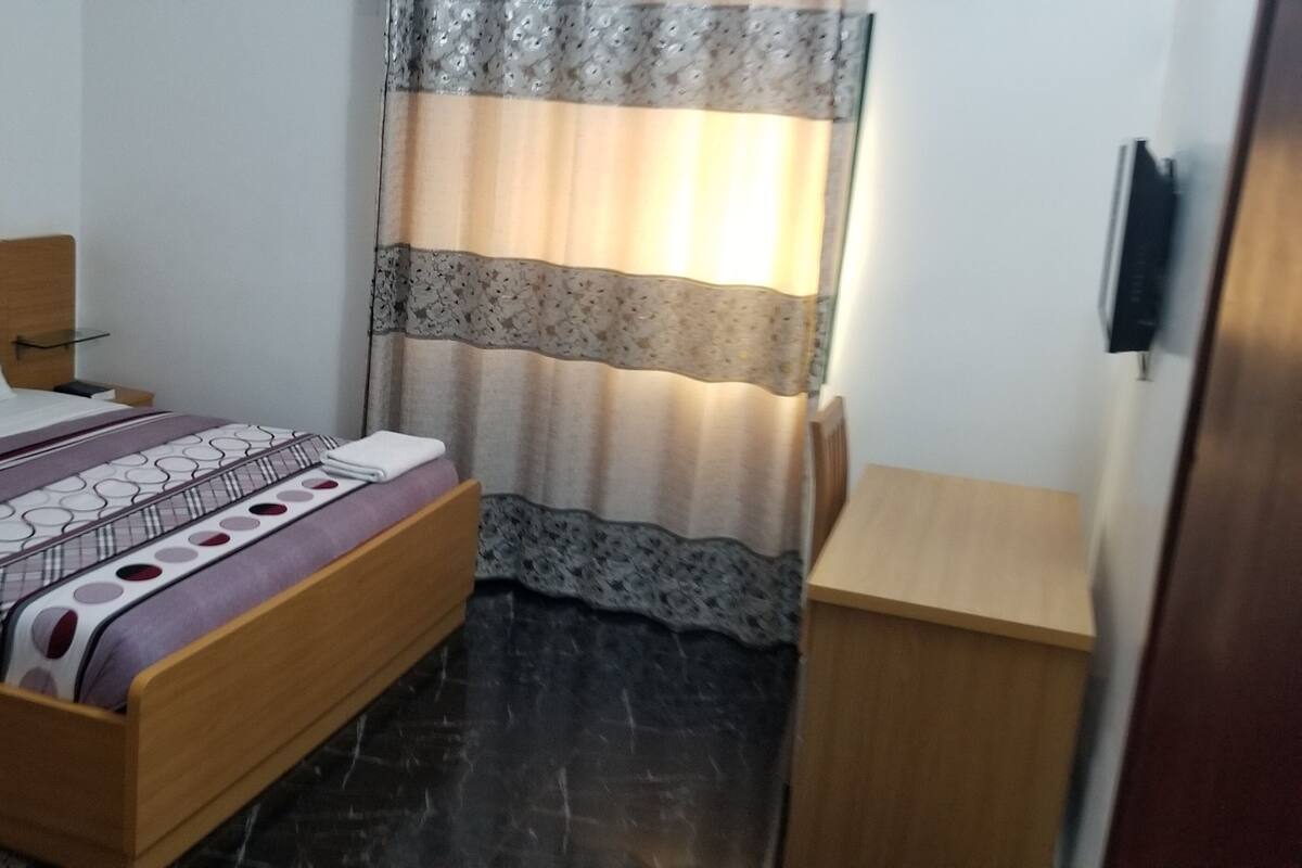 A bedroom with a wooden bed, a bedside table, a nightstand, a TV mounted on the wall, and a window with patterned curtains.