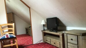 A small, compact room with a single bed, a wooden chair, a television on a wooden stand, and a small table with a lamp.