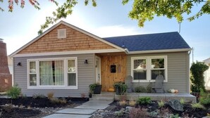 A single-story house with a grey siding exterior, a front porch with a wooden door, and a small garden area with various plants and a pathway leading to the door. - University Area Bungalow - The Moxie Harris House (Eugene)