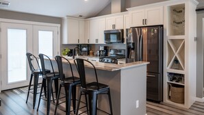 A modern kitchen with white cabinets, stainless steel appliances, and a central island with bar stools. - University Area Bungalow - The Moxie Harris House (Eugene)
