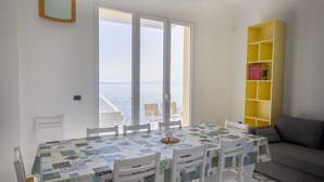 A dining area with a table and chairs, a yellow bookshelf, and a view of the sea through large windows. - Villa Francesca Sicily 8, Apartment in Villa on the sea (casteldaccia)
