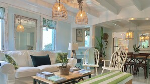Una sala de estar acogedora con un sofá blanco, un banco de madera y una mesita de centro. Hay dos lámparas colgantes, un espejo y una ventana con cortinas.