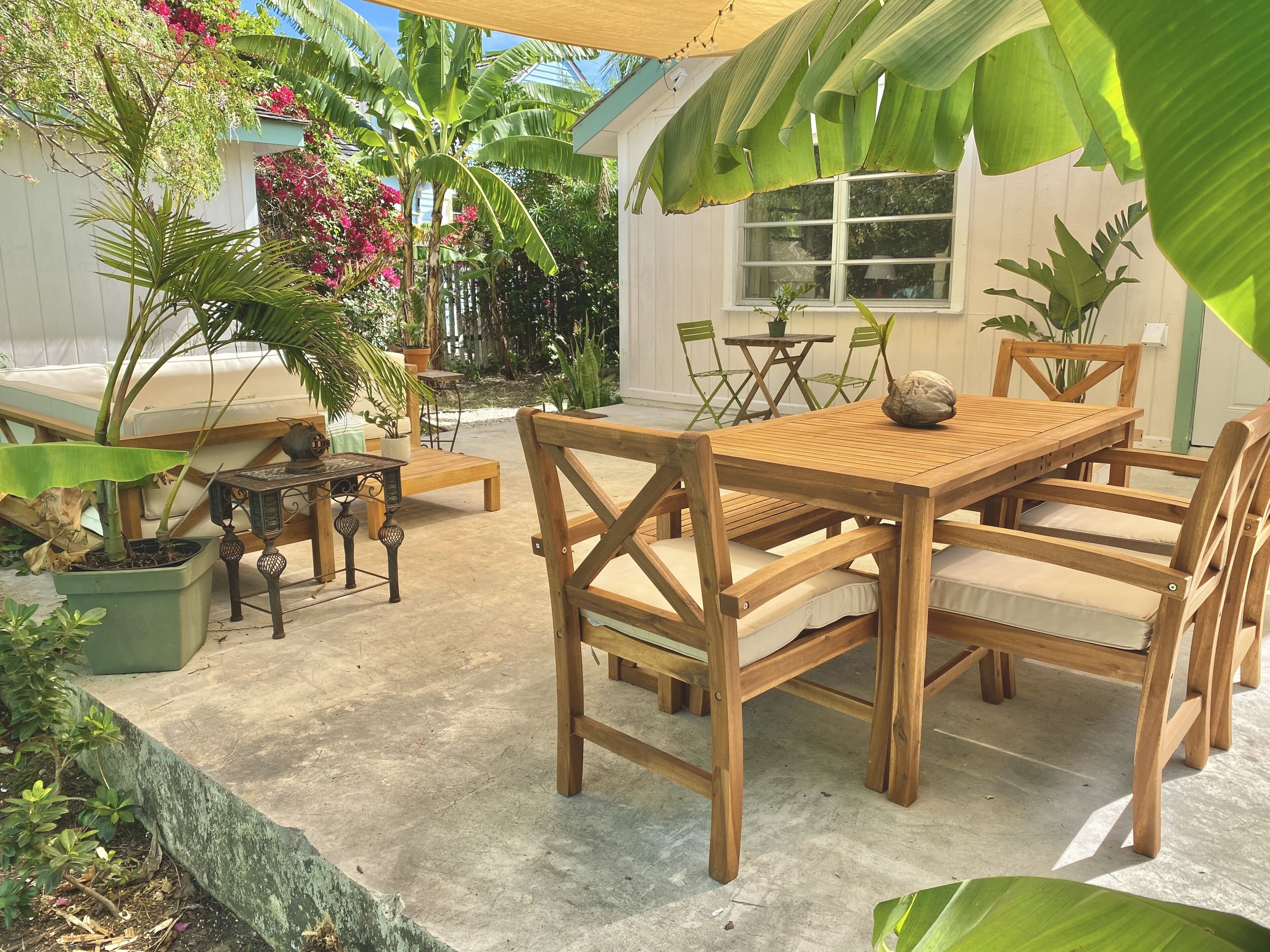 Un área de comedor al aire libre de madera con una mesa y sillas, rodeada de plantas en macetas y un edificio blanco al fondo.