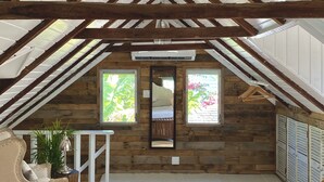 Un dormitorio en ático con techo inclinado, vigas de madera, dos ventanas con vistas a follaje, un espejo, una silla y una mesa.