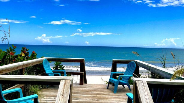 Eine Holzterrasse mit zwei blauen Stühlen, die zum Meer hinweisen.