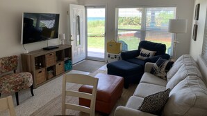 Ein Wohnzimmer mit Fernseher, Sofa, Hocker, Sessel und einer Tür, die auf einen Balkon führt.