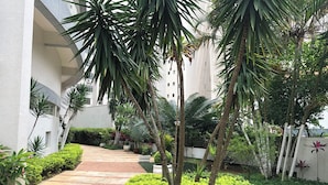 A pathway leading to a building with a white facade and a garden area with various plants and a palm tree. - High standard apt 200mts and sea view and complete recreation area (Guarujá)