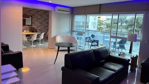 A modern living room with a black leather sofa, white chairs, a dining area with a brick wall, and large windows offering a view of the sea. - High standard apt 200mts and sea view and complete recreation area (Guarujá)