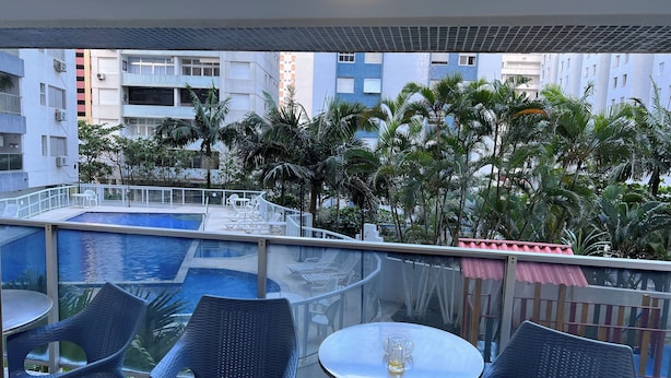 A rooftop pool area with lounge chairs and tables, surrounded by palm trees and buildings. - High standard apt 200mts and sea view and complete recreation area (Guarujá)