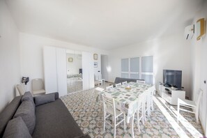 A modern living room with a gray sofa, a dining area with a patterned tablecloth, and a TV on a white stand.