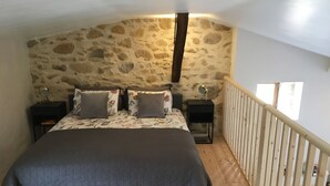 Un dormitorio con pared de piedra, suelo de madera, cama con colcha oscura, dos mesitas de noche con lámparas y barandilla de madera.