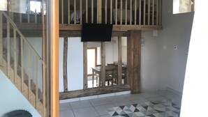 Una habitación con techo alto, una escalera de madera, un comedor con mesa y sillas, y un televisor colgado en la pared.