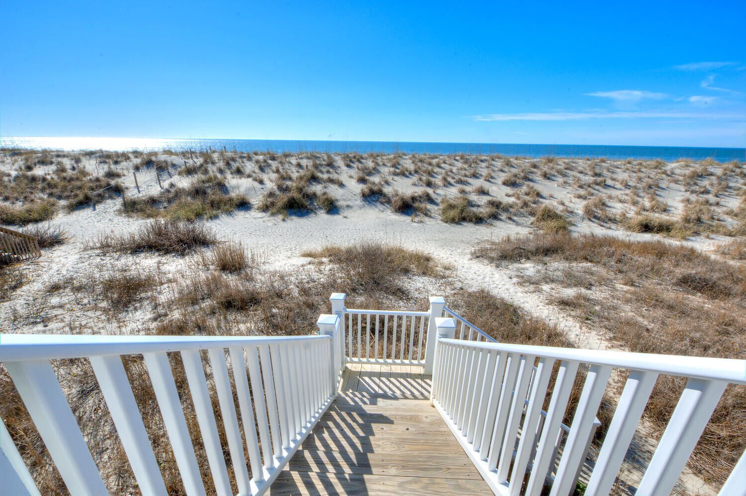 Une promenade en bois qui mène à une plage bordée de dunes, sous un ciel bleu et dégagé.