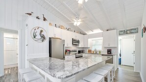 A spacious kitchen with white cabinetry, a large marble island with bar stools, and a ceiling fan. - 4BR 2BA OCEANFRONT COTTAGE- Fish, Fun & Sun! Relax, Recharge, Repeat!- Book Now! (Oak Island)