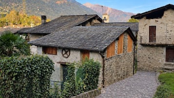 Ein Kopfsteinpflasterweg führt zu traditionellen Steinhäusern mit Holzhörnern, umgeben von Grünflächen und Bergen im Hintergrund.