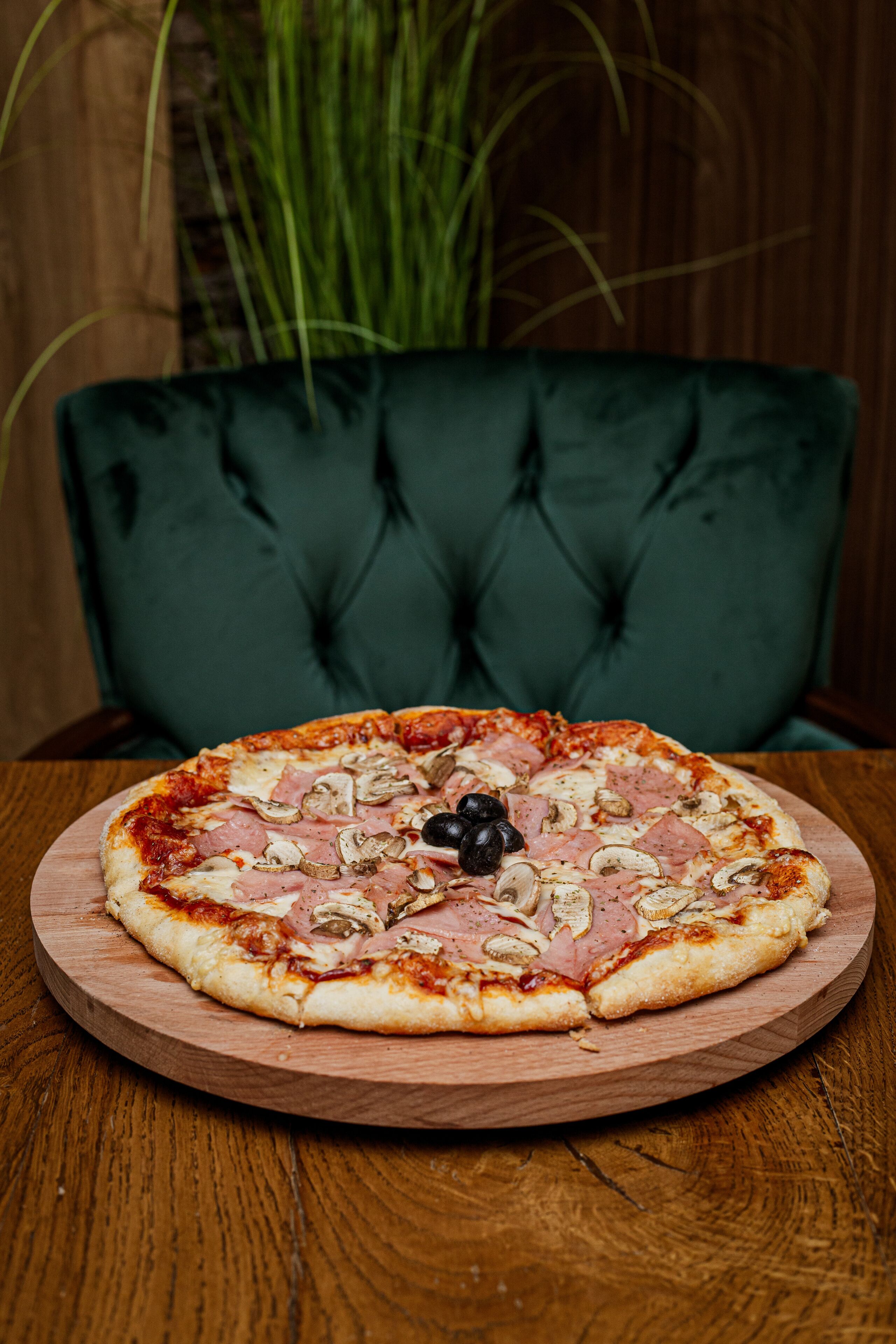 A pizza on a wooden board on a table with a green upholstered chair in the background.
