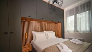 Une chambre à coucher moderne avec une tête de lit en bois, un grand lit recouvert d’une couette beige et une table de chevet dotée d’une télécommande.