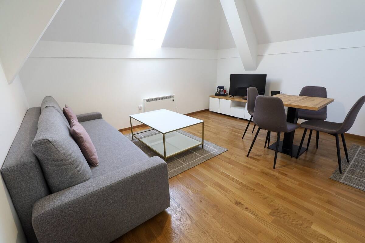 A modern loft apartment with a gray sofa, a glass coffee table, a dining table with chairs, a TV, and a radiator.