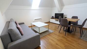 Un loft moderno con sofá gris, una mesa de centro de vidrio, una mesa de comedor con sillas, televisor y radiador.