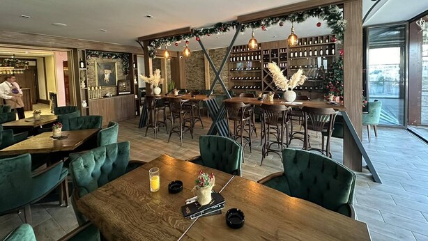 Un restaurant avec des tables en bois et des chaises à l’assise rembourrée de tissu vert, un coin bar décoré d’un sapin de Noël et, au fond, un meuble à vin.