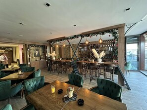 Un restaurant avec des tables en bois et des chaises à l’assise rembourrée de tissu vert, un coin bar décoré d’un sapin de Noël et, au fond, un meuble à vin.