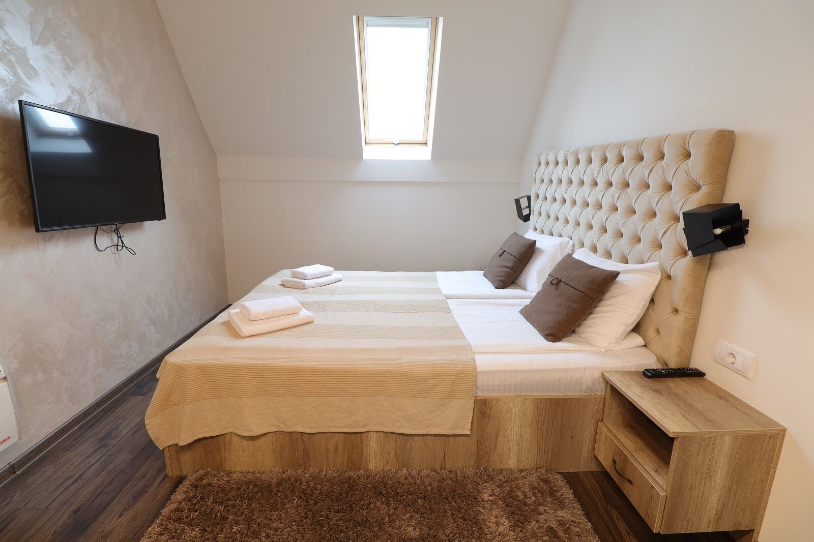 A bedroom with a large bed, a TV mounted on the wall, a window, and a wooden nightstand.