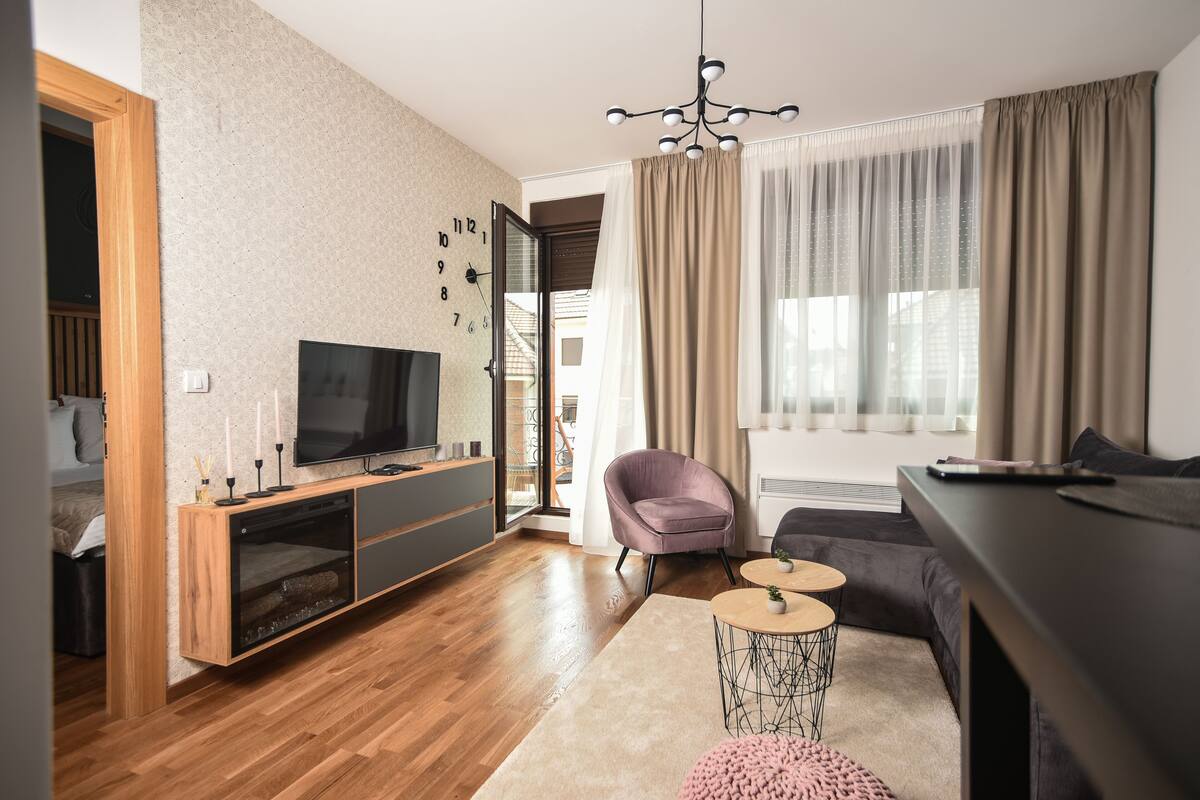 A modern living room with a wooden floor, a TV on a wooden stand, a purple armchair, a sofa, a coffee table, and a large window with curtains.