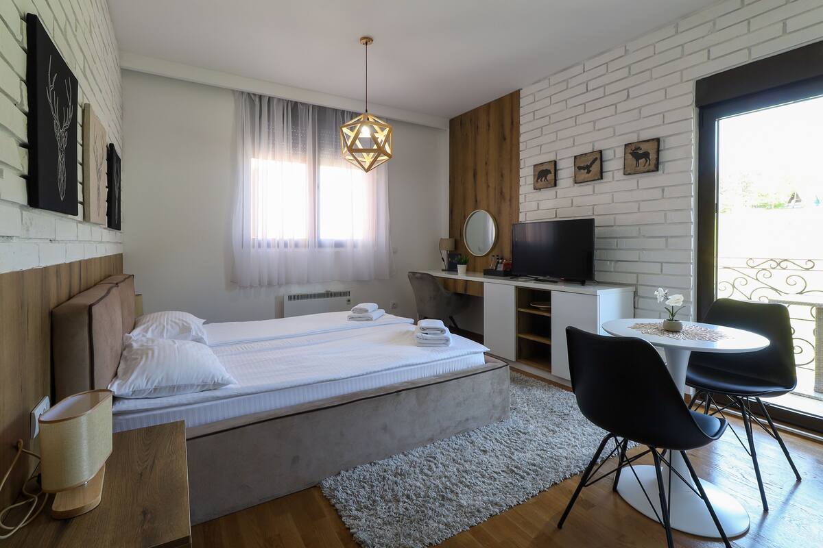 A modern bedroom with a large bed, a desk, a chair, a TV, and a balcony with a railing.
