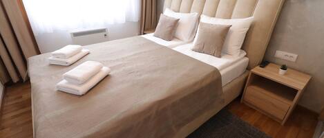 Una cama bien hecha con colcha y almohadas color beige, una mesita de noche con lámpara y una ventana con cortinas.