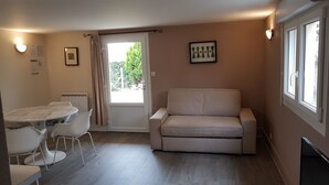 Een kleine, goed verlichte kamer met een witte bank, een eettafel met stoelen en een raam met gordijnen.