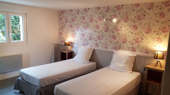 Une chambre avec deux lits simples, un papier peint à motifs floraux, des lampes de chevet et une fenêtre avec des stores.