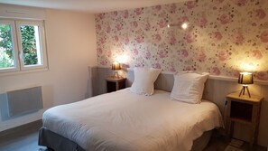 Une chambre à coucher avec un grand lit, un papier peint à motifs floraux, deux lampes de chevet et une fenêtre avec des stores.