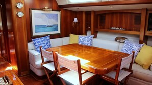 Interior de un yate con comedor que incluye una mesa y sillas de madera, un sofá con cojines acolchados y estampados, un cuadro enmarcado de un velero y un pequeño estante con objetos decorativos.