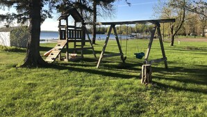 Un parque infantil con un columpio de madera y tobogán, rodeado de césped y árboles, cerca de un cuerpo de agua.