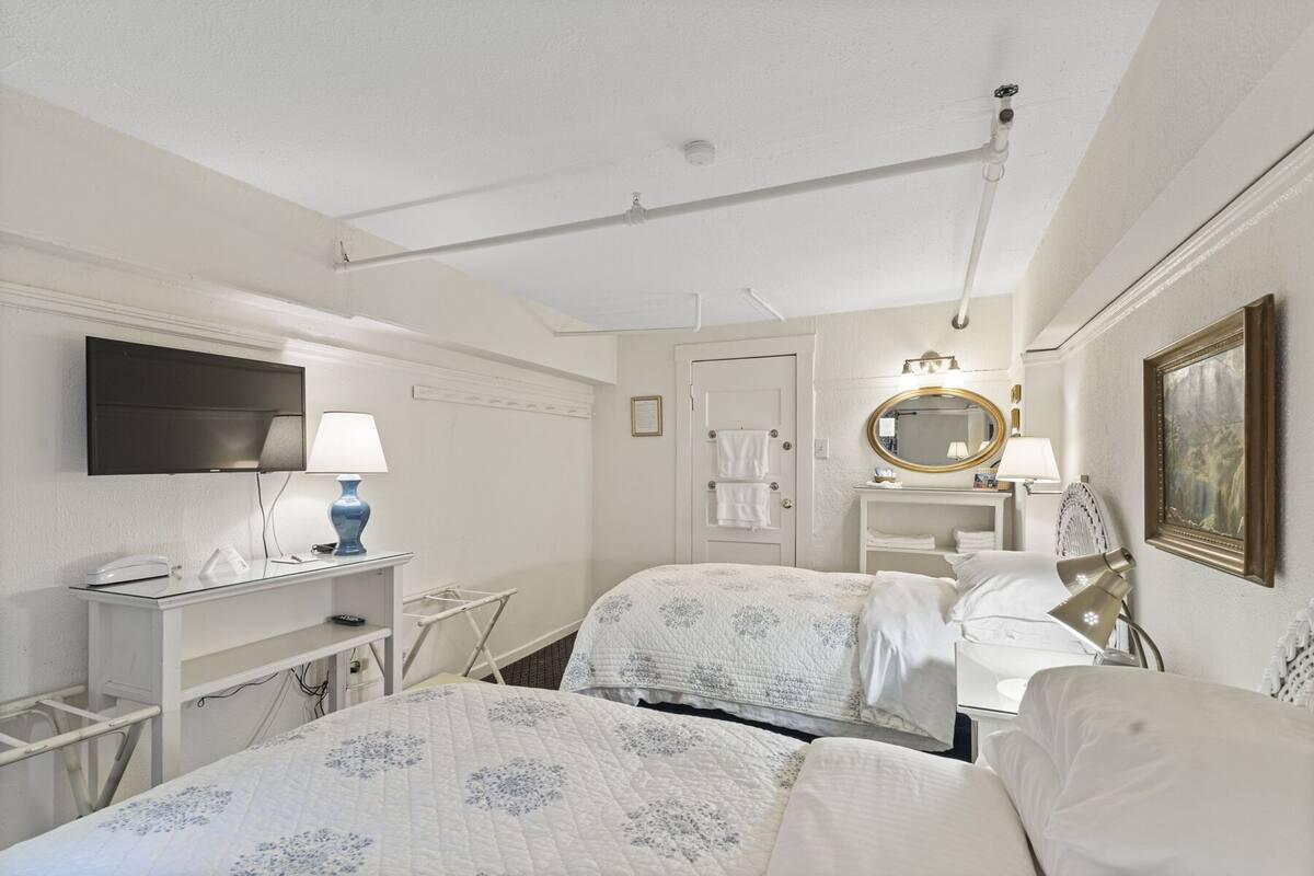 A hotel room with two beds, a TV, a desk with a lamp, a bathroom with a mirror, and a painting on the wall.