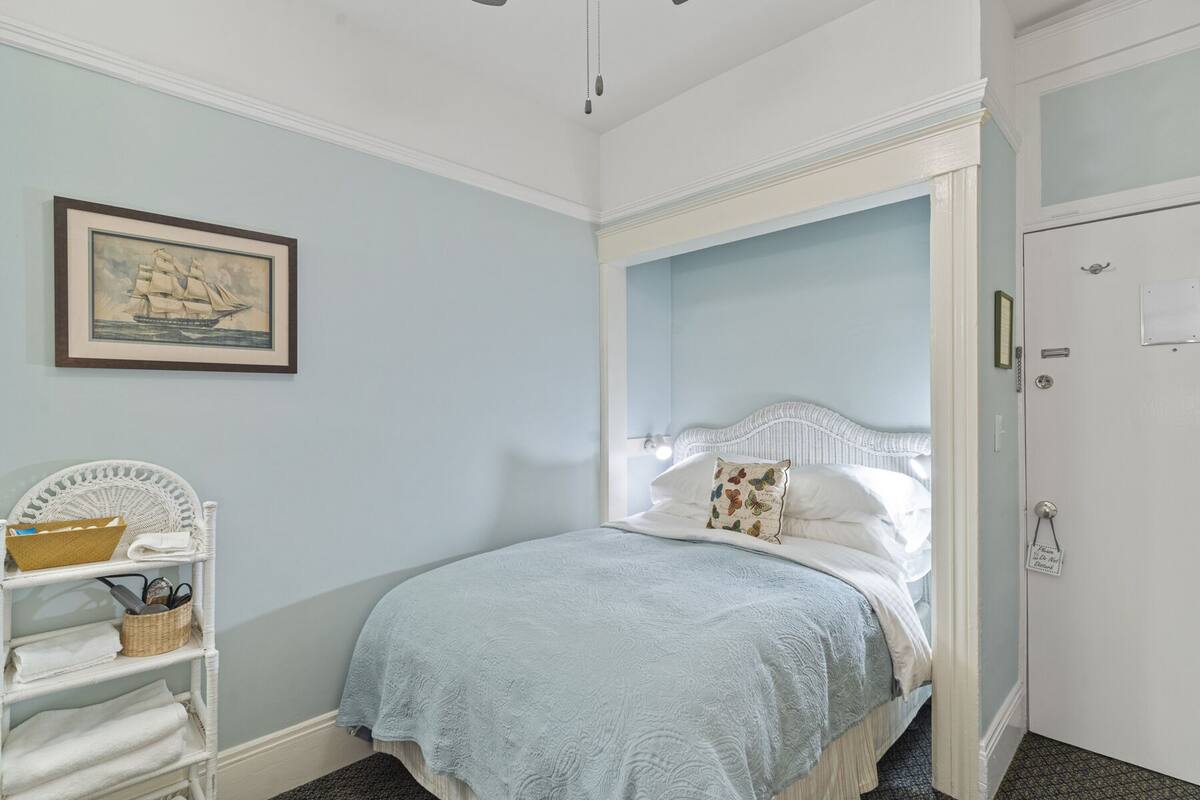 A bedroom with a bed, a white wicker chair, a shelf with towels, and a framed picture on the wall.