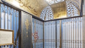 A vintage elevator interior with ornate metal railings, a control panel, and decorative ceiling elements.
