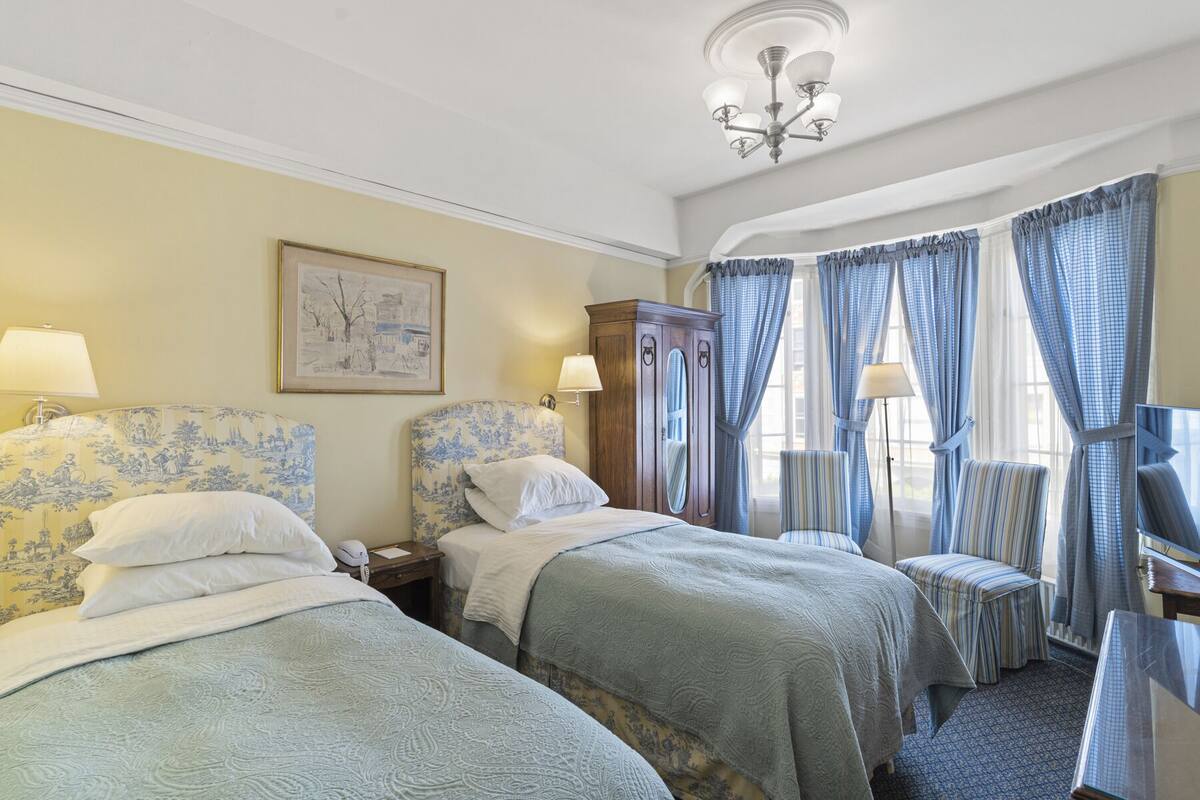 A hotel room with two beds, a wooden wardrobe, a painting on the wall, and a chandelier.