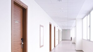 Un long couloir bien éclairé, avec des portes en bois et des murs blancs, qui mène à une pièce où se trouve une enseigne de sortie verte.