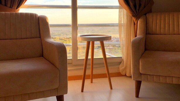 Une pièce avec deux fauteuils placés face à une grande fenêtre ornée de rideaux, une petite table ronde et une vue sur un paysage lointain.