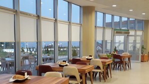 Un vaste espace intérieur aménagé en salle à manger, doté de grandes fenêtres offrant une vue sur l’extérieur. Les tables sont disposées en rangées, accompagnées de chaises.