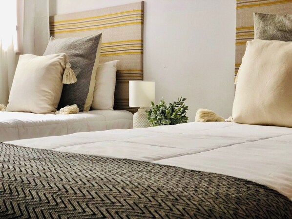 Habitación de hotel con dos camas, cabecero a rayas horizontales y una mesita de noche pequeña con una lámpara y una planta.