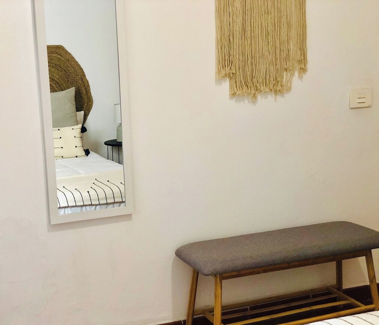 A bedroom with a bed, a bench, a mirror, and a wall-mounted macrame piece.