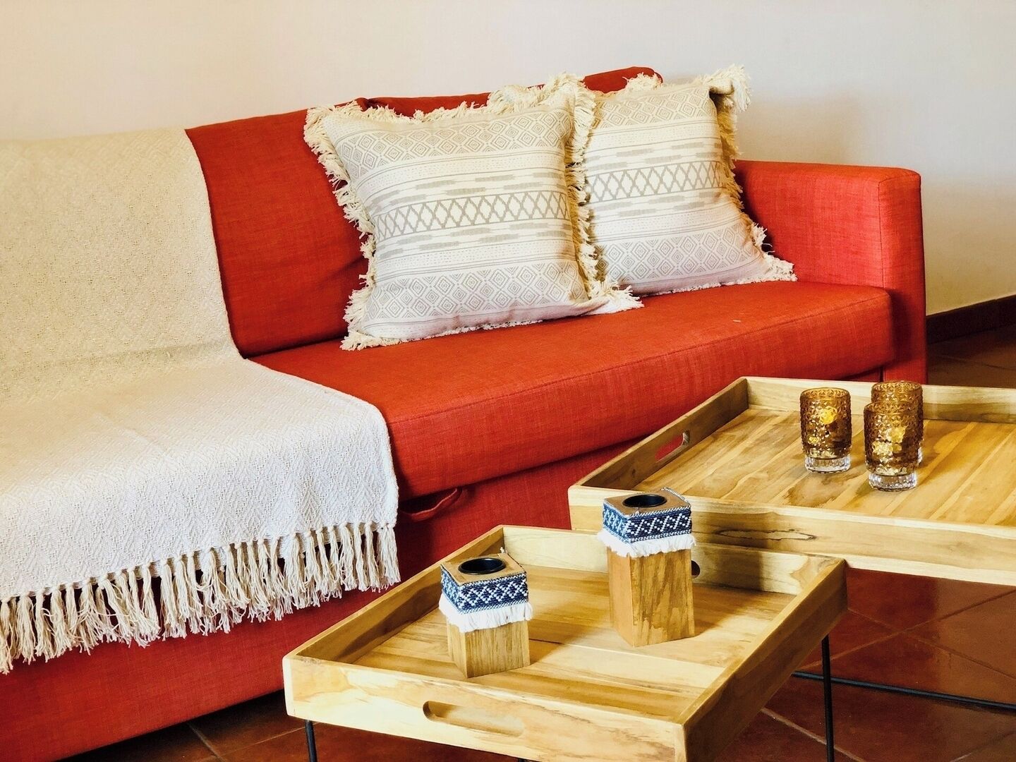 A red sofa with white and patterned cushions, a wooden tray with two glass containers, and a white throw blanket.