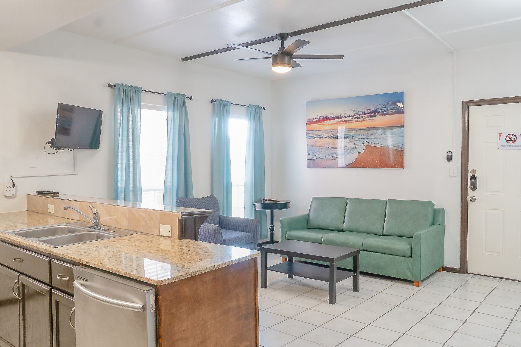 A compact living space with a kitchenette, a green sofa, a flat-screen TV, and a painting of a beach scene.